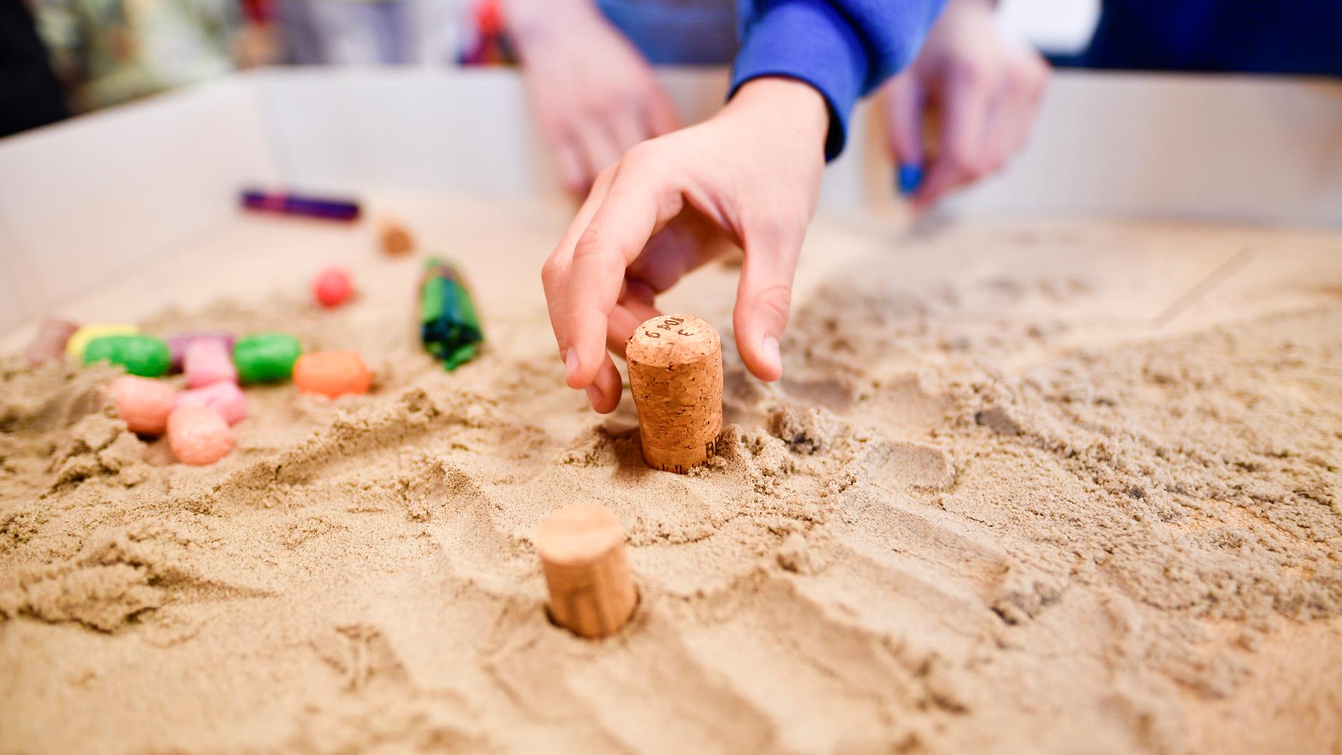 Eine Kinderhand greift nach einem Korken, der in einem Sandkasten steckt. Es handelt sich um den ersten Inklusionsspielplatz für Schulkinder. 