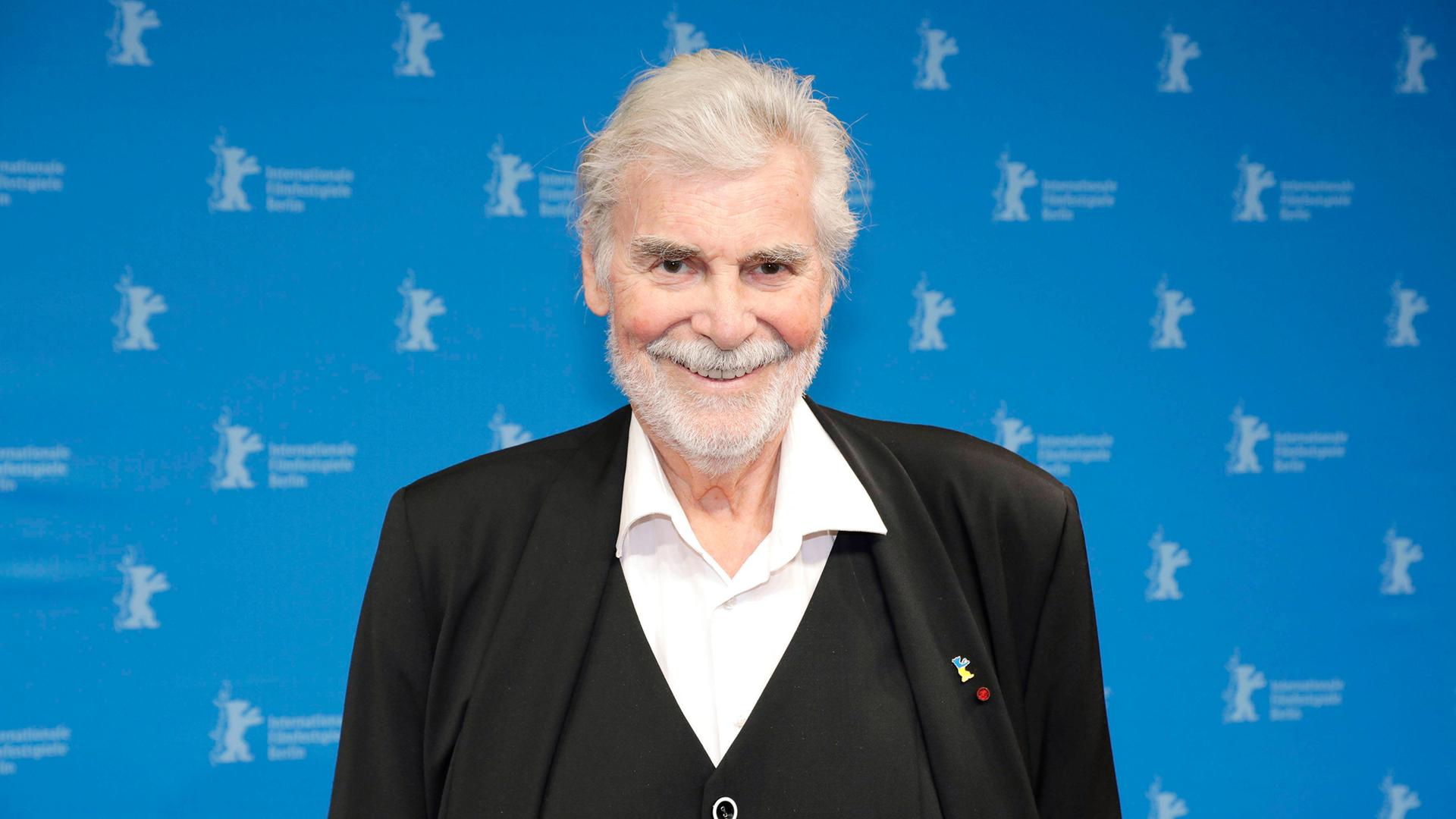 Peter Simonischek steht im schwarzen Anzug vor einem blauen Hintergrund mit kleinen Berlinale Logos.