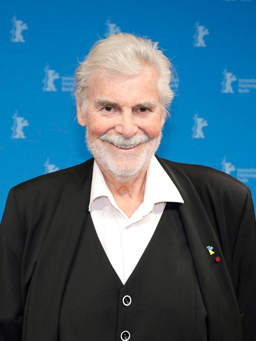 Peter Simonischek steht im schwarzen Anzug vor einem blauen Hintergrund mit kleinen Berlinale Logos.