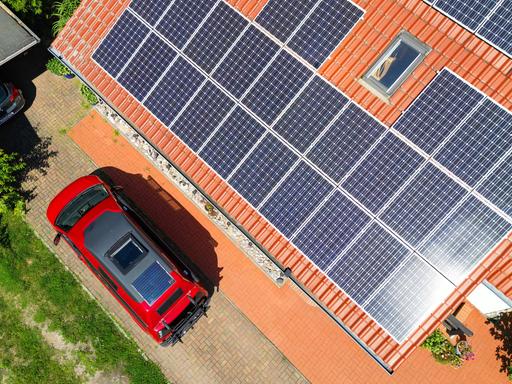 Eine großflächige Photovoltaikanlage befindet sich auf einem rot gedeckten Hausdach (Luftaufnahme mit einer Drohne). Davor steht ein roter Camper-Van mit einem integrierten Solarmodul auf dem Fahrzeugdach. Eine großflächige Photovoltaikanlage befindet sich auf einem rot gedeckten Hausdach (Luftaufnahme mit einer Drohne). Davor steht ein roter Camper-Van mit einem integrierten Solarmodul auf dem Fahrzeugdach.