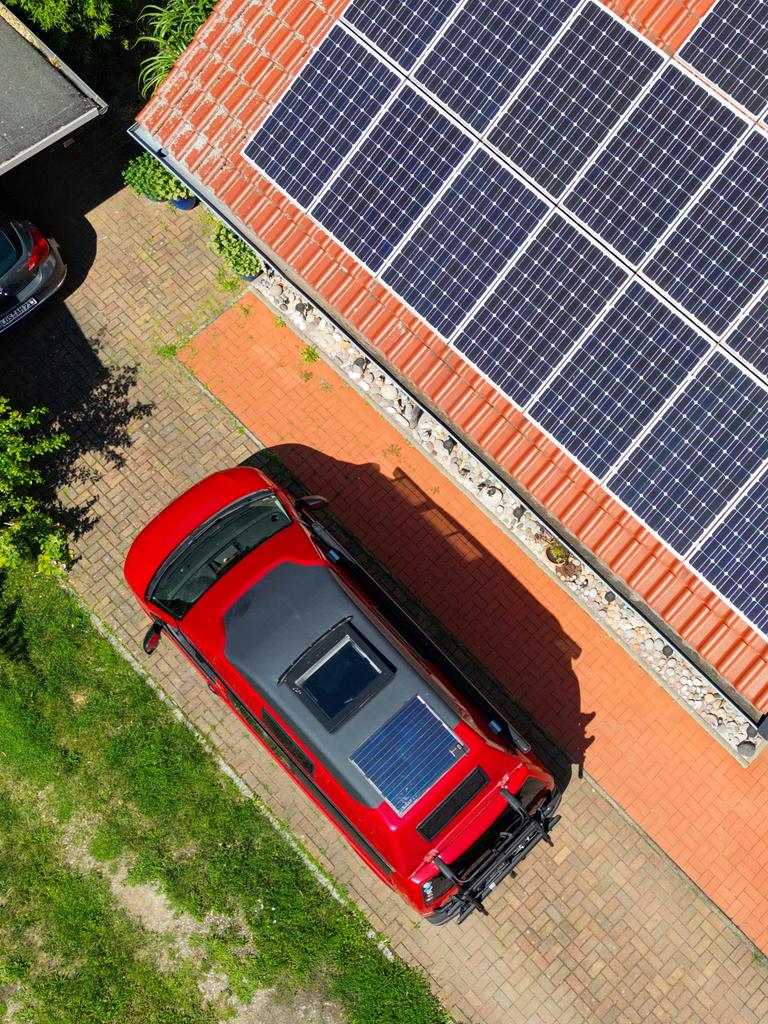 Eine großflächige Photovoltaikanlage befindet sich auf einem rot gedeckten Hausdach (Luftaufnahme mit einer Drohne). Davor steht ein roter Camper-Van mit einem integrierten Solarmodul auf dem Fahrzeugdach. Eine großflächige Photovoltaikanlage befindet sich auf einem rot gedeckten Hausdach (Luftaufnahme mit einer Drohne). Davor steht ein roter Camper-Van mit einem integrierten Solarmodul auf dem Fahrzeugdach.