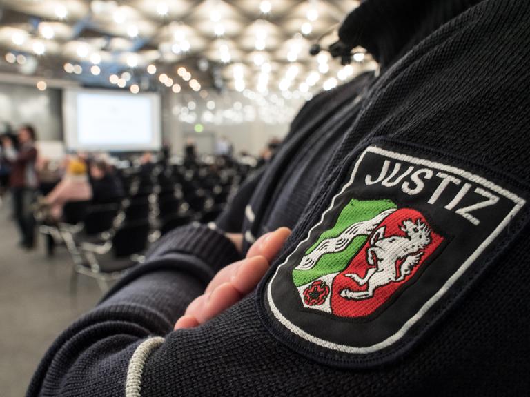 Blick auf das Justiz-Symbol auf dem Ärmel eines Justiz-Mitarbeiters in Düsseldorf. Im Hintergrund: Blick in den Saal. 