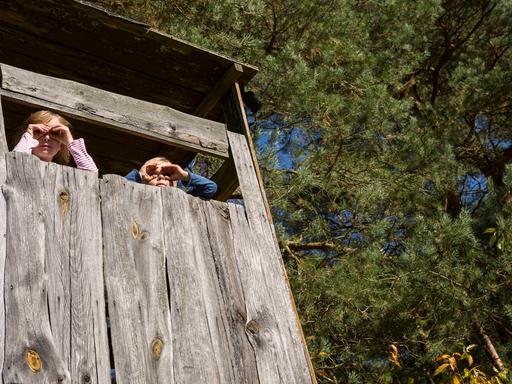 Ein junge und ein Mädchen schauen aus einem Baumhaus und halten sich die Hände wir Ferngläser vor die Augen.