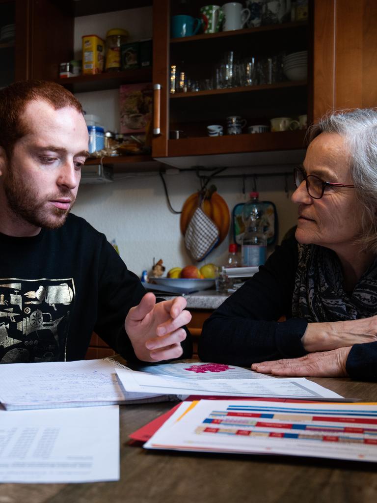 Eine ältere Frau und ein junger Mann sitzen nebeneinander am Küchentisch und sehen sich Unterlagen zum Sprachenlernen an.