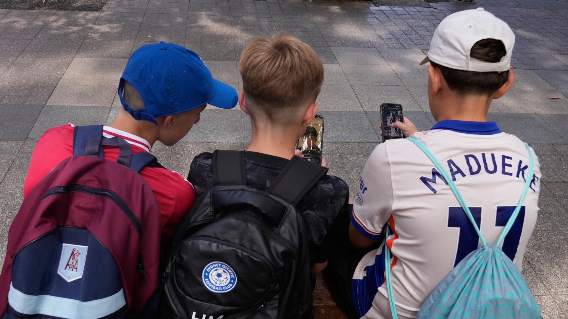 Drei Elfjährige benutzen ihre Handys, während sie vor einer Schule in Sydney sitzen. Man sieht die Jungen von hinten. 