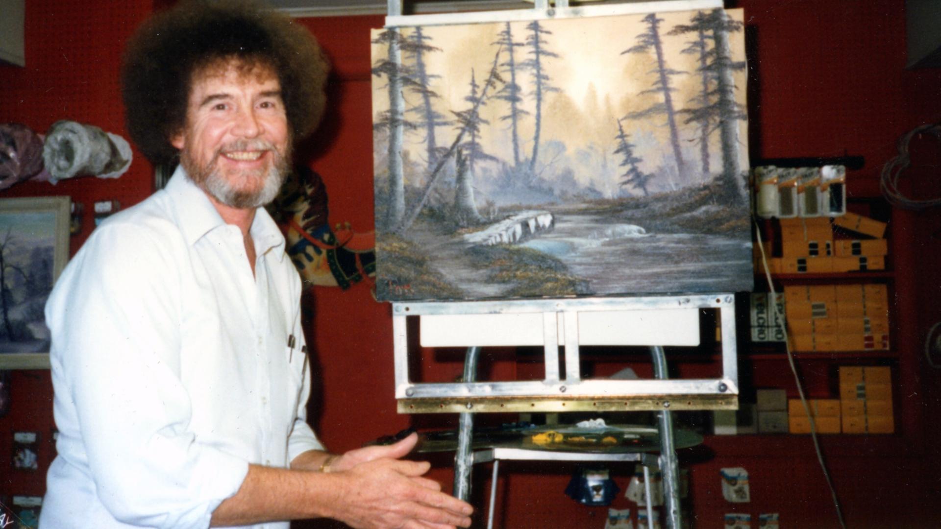 Bob Ross neben einem Landschafts-Gemälde von ihm auf einer Staffelei.