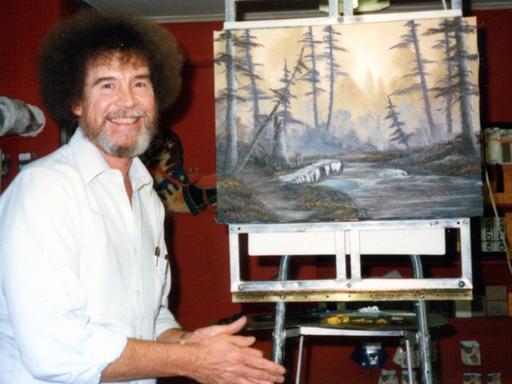 Bob Ross neben einem Landschafts-Gemälde von ihm auf einer Staffelei.