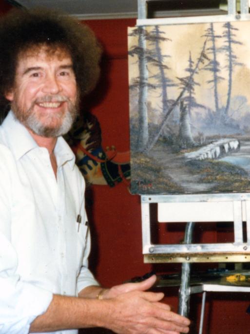 Bob Ross neben einem Landschafts-Gemälde von ihm auf einer Staffelei.