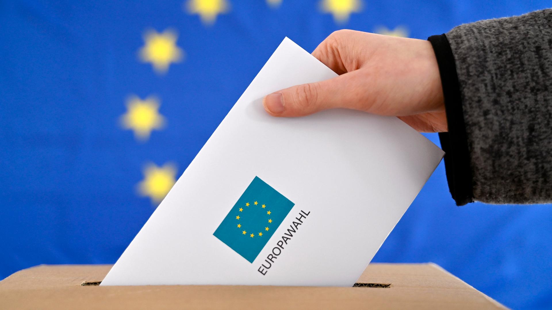 Ein Wahl-Brief wird in eine Wahl-Urne gesteckt. Im Hinter-Grund ist eine EU-Flagge. 