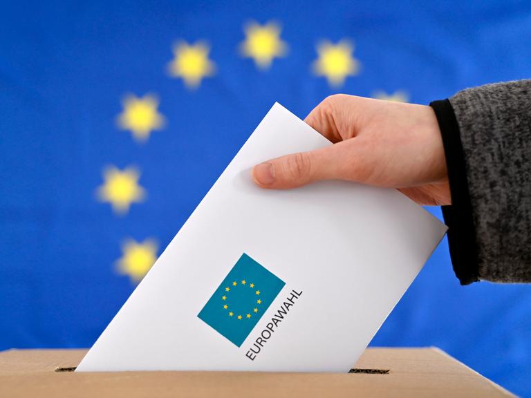 Ein Wahlbrief wird in eine Wahlurne gesteckt, im Hintergrund eine EU-Flagge. 