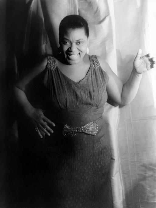 Die afroamerikanische Bluessängerin Bessie Smith in einem schwarzweißen Porträtm am 3. Februar 1936. Die Sängerin trägt ein langes Kleid, kurze Haare und lächelt.