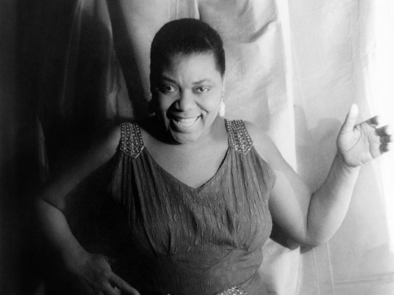 Die afroamerikanische Bluessängerin Bessie Smith in einem schwarzweißen Porträtm am 3. Februar 1936. Die Sängerin trägt ein langes Kleid, kurze Haare und lächelt.