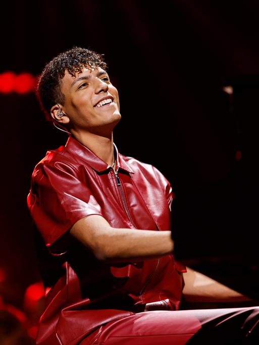 Louis Philippson sitzt bei einem Auftritt lächelnd am Klavier. Er hat dunkles, gelocktes Haar und trägt ein burgunderrotes Lederhemd.