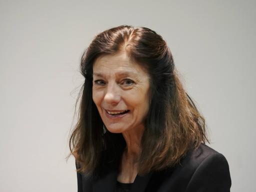Die Autorin Ursula Krechel auf einem Porträtfoto. Sie lächelt verhalten und schaut leicht seitlich. Der Kopf ist leicht gebeugt, das braune Haar offen. Sie steht vor einem grauen Hintergrund. 
