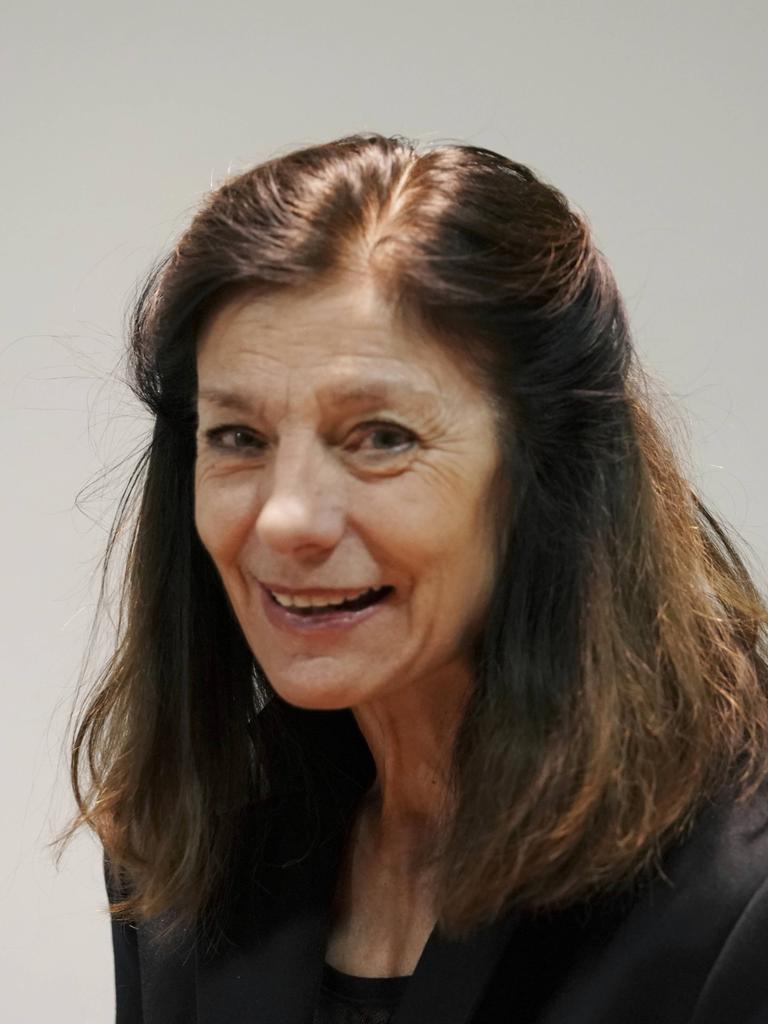 Die Autorin Ursula Krechel auf einem Porträtfoto. Sie lächelt verhalten und schaut leicht seitlich. Der Kopf ist leicht gebeugt, das braune Haar offen. Sie steht vor einem grauen Hintergrund. 