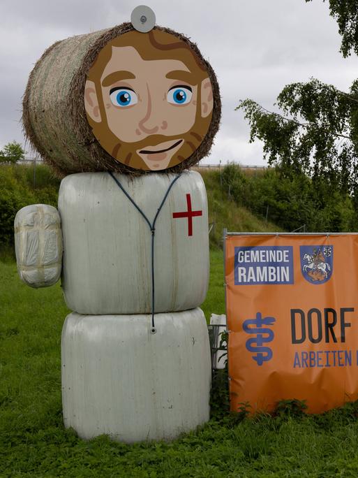 Eine Strohpuppe, die aussieht wie ein Arzt, steht neben einem Schild an einer Straße - darauf steht: "Dorf sucht Arzt".