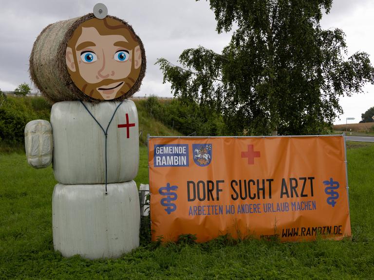 Eine Strohpuppe, die aussieht wie ein Arzt, steht neben einem Schild an einer Straße - darauf steht: "Dorf sucht Arzt".