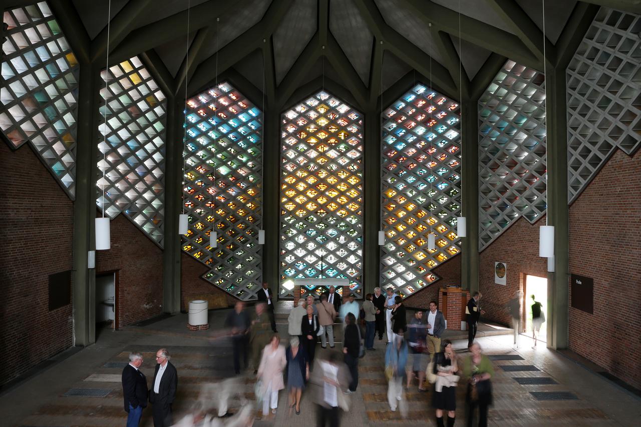 In der Eingangshalle der Hamburger Kapernaum-Kirche, sind Menschen zu einer Denkmal-Führung versammelt. Durch kleine, bunte Glasfenster, die in einem Bienenwabenmuster aus Beton angeordnet sind, fällt Licht in die Halle.
