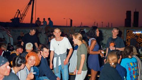 Zu sehen ist ein Gruppe von Frauen und Männer auf einer Party im Freien. Im Hintergrund rötlicher gefärbter Himmel vor urbaner Stadtlandschaft.