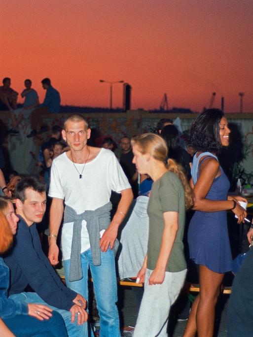 Zu sehen ist ein Gruppe von Frauen und Männer auf einer Party im Freien. Im Hintergrund rötlicher gefärbter Himmel vor urbaner Stadtlandschaft.