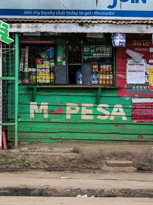 Zwei Fußgänger laufen in Kenia an einem Kiosk vorbei, auf dem für M-Pesa geworben wird.