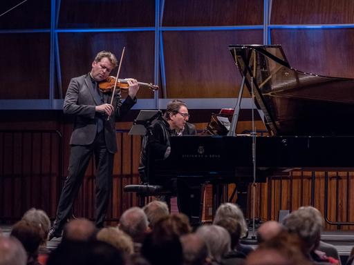 Ein Geiger und ein Pianist geben ein Konzert in einem voll besetzten, holzvertäfelten Saal, der blau beleuchtet ist.