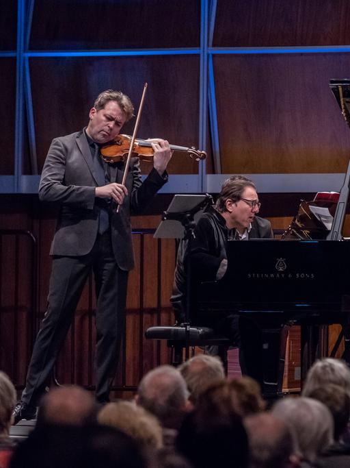 Ein Geiger und ein Pianist geben ein Konzert in einem voll besetzten, holzvertäfelten Saal, der blau beleuchtet ist.
