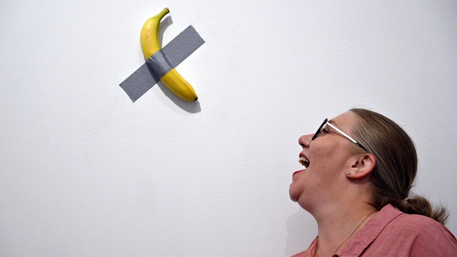 Eine Banane ist mit Panzertape an einer weißen Wand befestigt. Eine Frau steht daneben und öffnet den Mund so, als ob sie die Banane essen wollte Eine Banane ist mit Panzertape an einer weißen Wand befestigt. Eine Frau steht daneben und öffnet den Mund so, als ob sie die Banane essen wollte