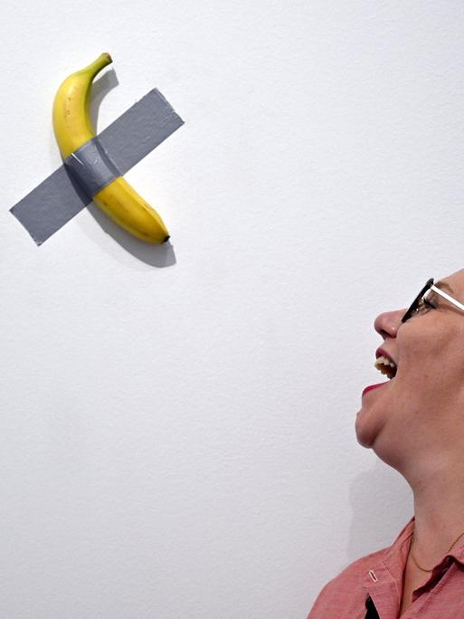 Eine Banane ist mit Panzertape an einer weißen Wand befestigt. Eine Frau steht daneben und öffnet den Mund so, als ob sie die Banane essen wollte