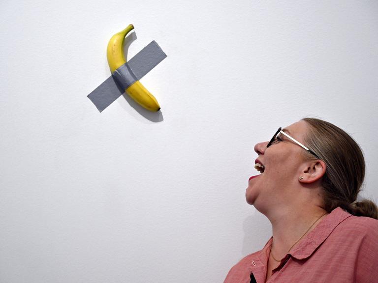 Eine Banane ist mit Panzertape an einer weißen Wand befestigt. Eine Frau steht daneben und öffnet den Mund so, als ob sie die Banane essen wollte