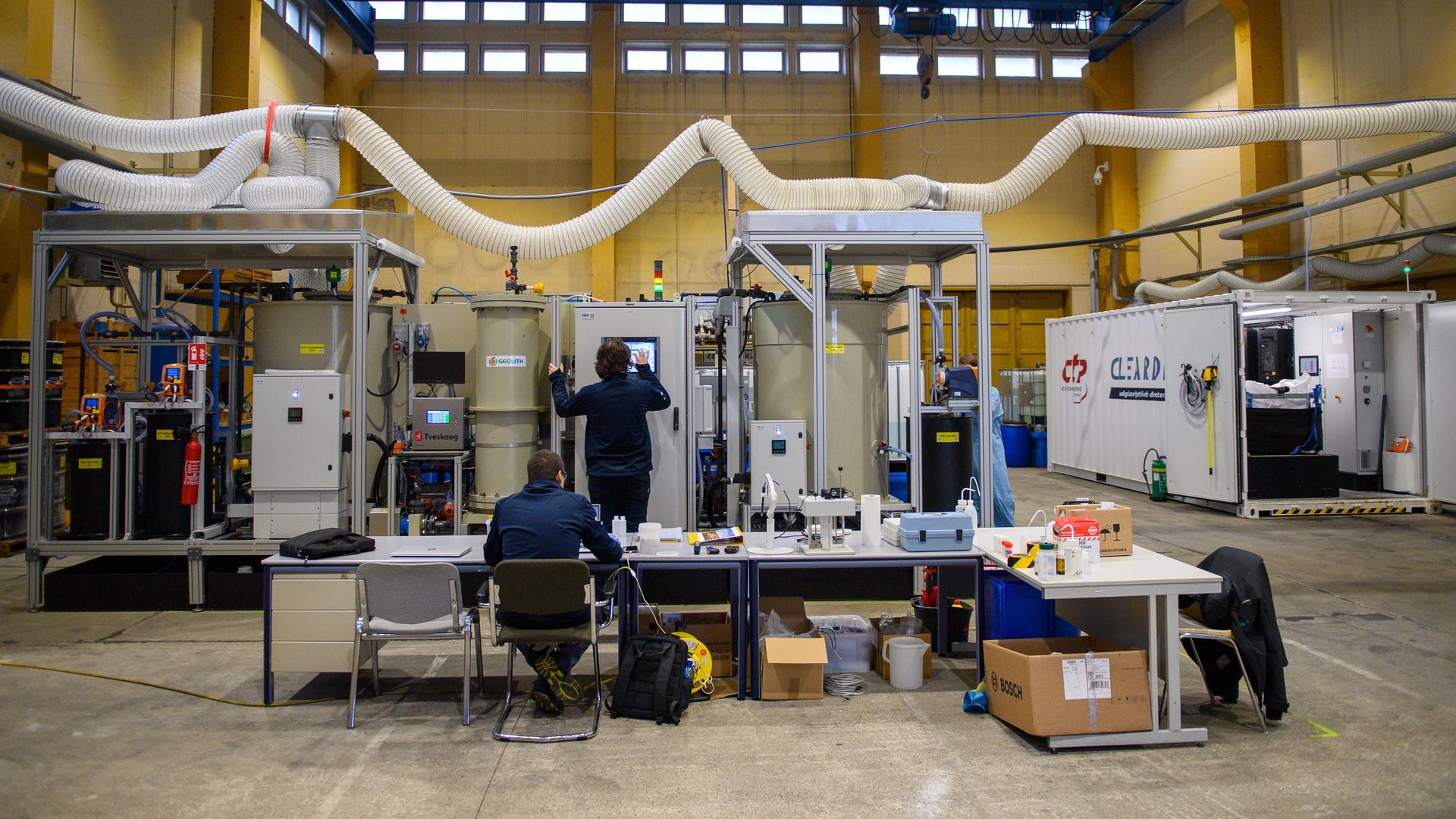 Techniker bedienen in einer Halle eine Apparatur mit vielen Schläuchen. 
