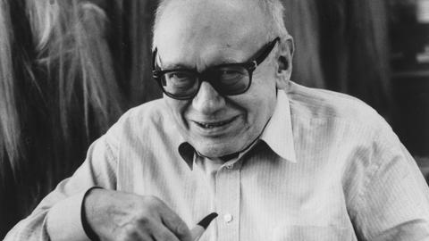 Auf dem schwarz-weiß Foto ist Ernst Jandl während einer Lesung im Theater Neumarkt in Zuerich im  Mai 1995 zu sehen. Er schaut in die Kamera. In der rechten Hand hält er eine Pfeife.