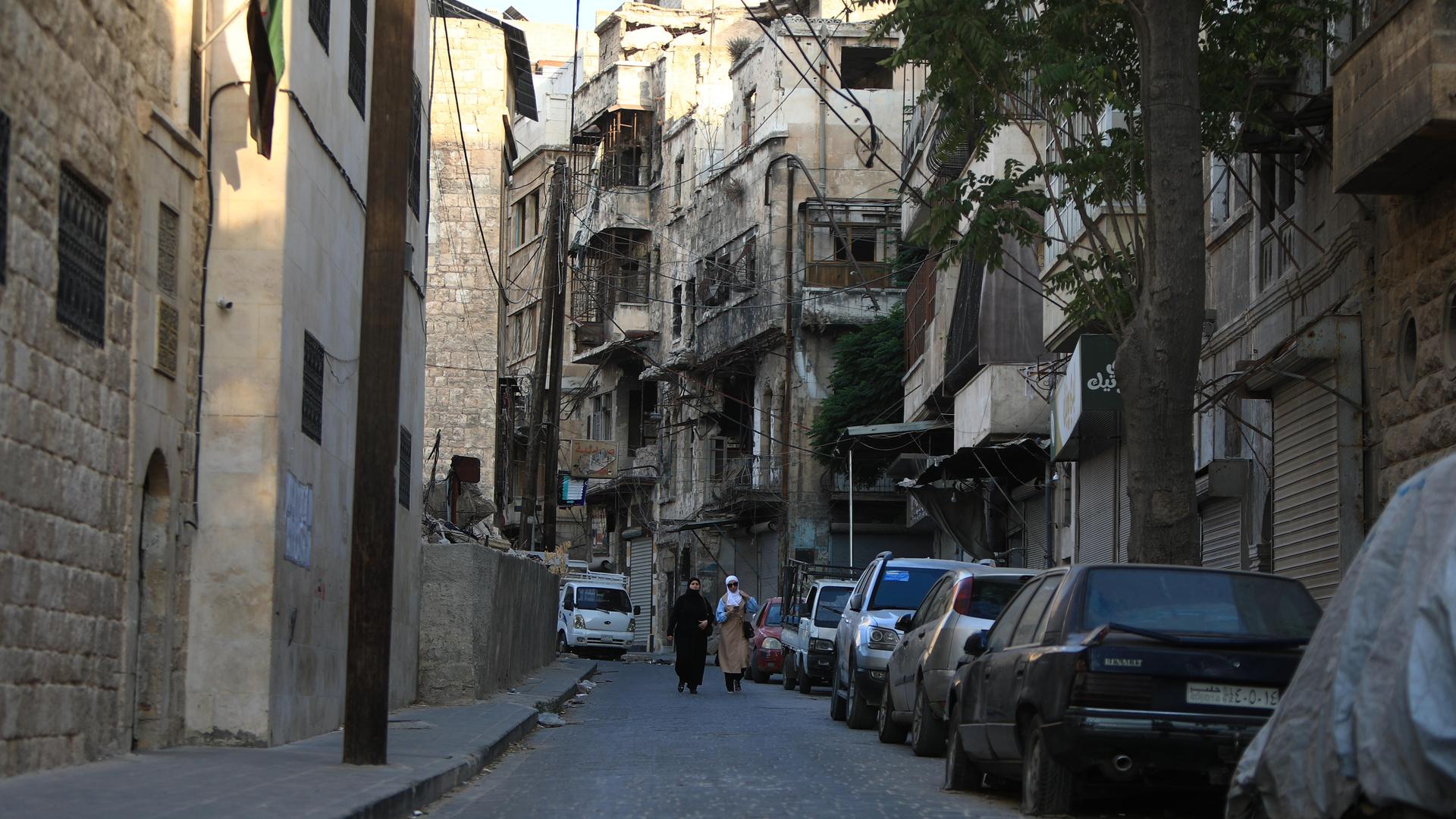 Auf einer Straße in Aleppo gehen zwei Menschen zwischen den Häusern an geparkten Autos vorüber. Auf einer Straße in Aleppo gehen zwei Menschen zwischen den Häusern an geparkten Autos vorüber.