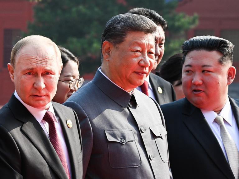 Das Bild zeigt Chinas Staatschef Xi Jinping in einem dunklen Anzug, der der Kleidung von Mao Tsetung nachempfunden ist. Neben ihm steht Russlands Präsident Putin und Machthaber Kim Jong Un aus Nordkorea. 