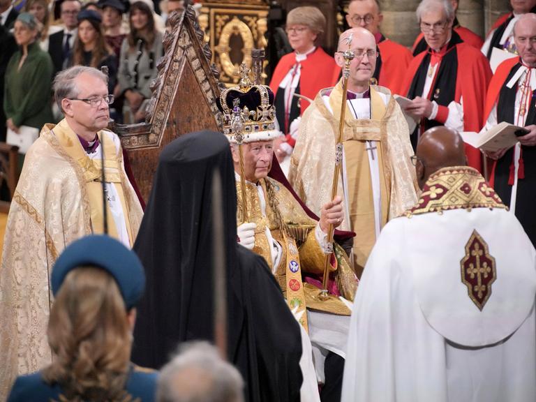 Der frisch gekrönte König Charles III. sitzt in der Westminster Abbey mit der Krone auf dem Haupt, in den Händen hält er das Zepter und den Stab.