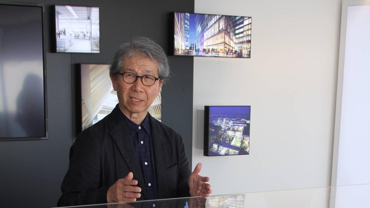 Der Architekt Riken Yamamoto steht hinter einem Glaskasten mit einem Architekturmodell, hinter ihm hängen Bilder von Bauwerken an der Wand.