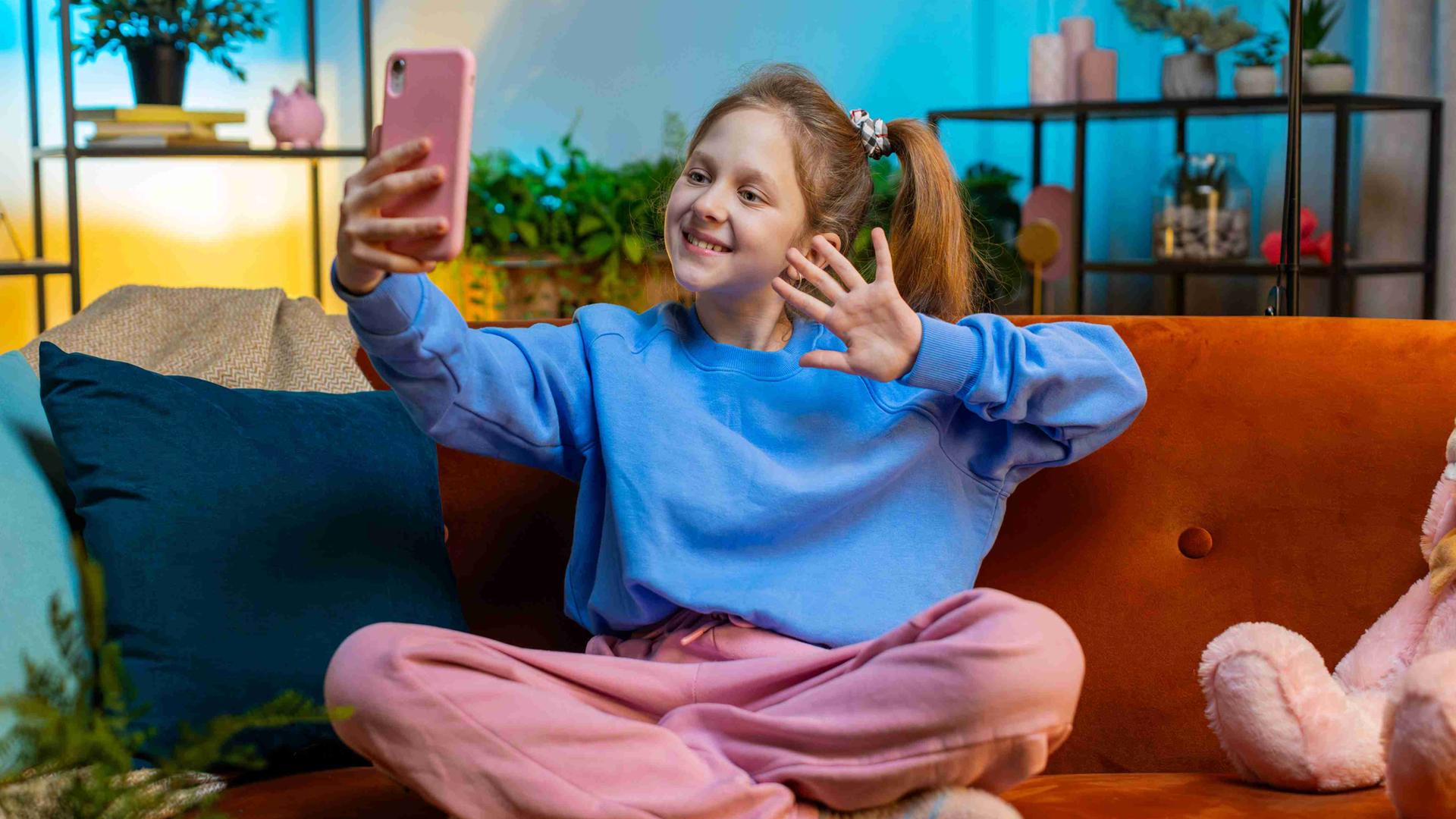 Kleine Influencerin: Ein Mädchen sitzt in pastellfarbener Kleidung in einem in Pastellfarben gehaltenen Wohnzimmer und winkt in ihr rosa Smartphone.