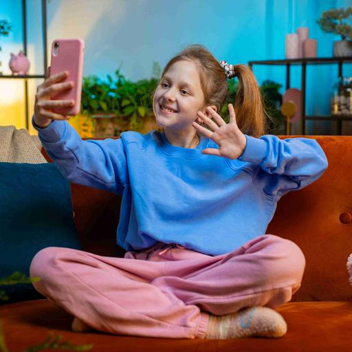 Kleine Influencerin: Ein Mädchen sitzt in pastellfarbener Kleidung in einem in Pastellfarben gehaltenen Wohnzimmer und winkt in ihr rosa Smartphone.