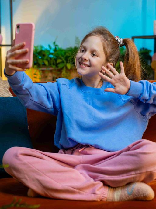 Kleine Influencerin: Ein Mädchen sitzt in pastellfarbener Kleidung in einem in Pastellfarben gehaltenen Wohnzimmer und winkt in ihr rosa Smartphone.