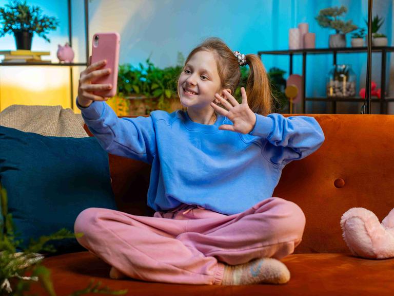 Kleine Influencerin: Ein Mädchen sitzt in pastellfarbener Kleidung in einem in Pastellfarben gehaltenen Wohnzimmer und winkt in ihr rosa Smartphone.