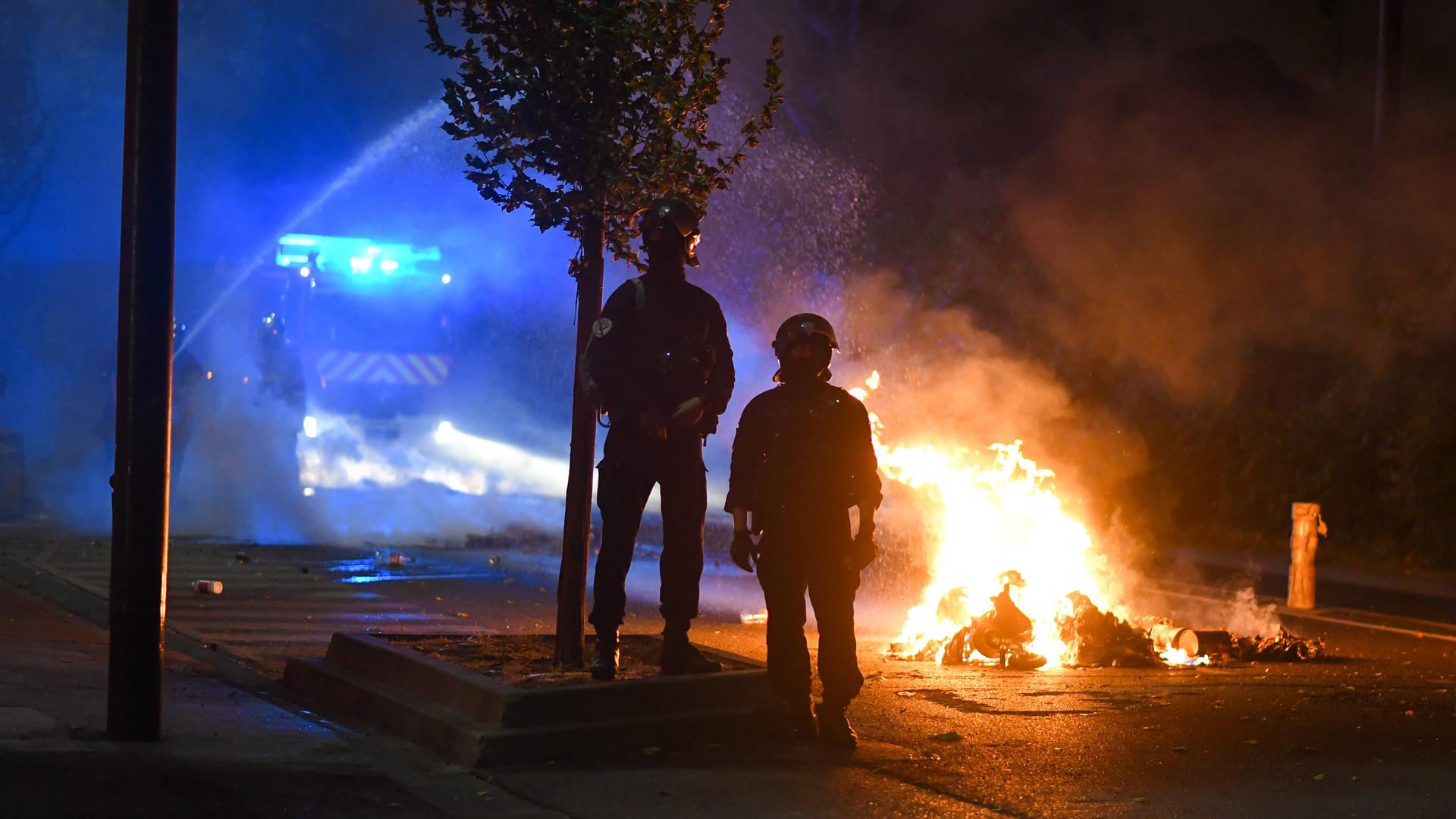 Feuerwehrmänner stehen in der Nacht zum 1. Juli auf der Straße vor brennenden Autos. Im Hintzergrund steht ein Löschwagen. 