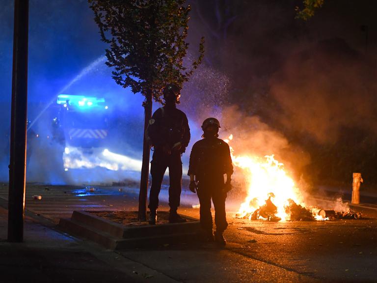 Feuerwehrmänner stehen in der Nacht zum 1. Juli auf der Straße vor brennenden Autos. Im Hintzergrund steht ein Löschwagen. 