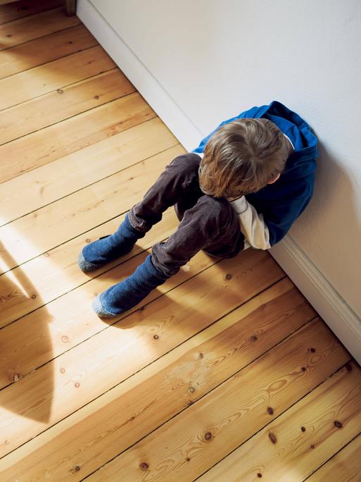  Ein Junge sitzt traurig in seinem Kinderzimmer neben seinem Schulranzen und versteckt sein Gesicht hinter seinen Händen.