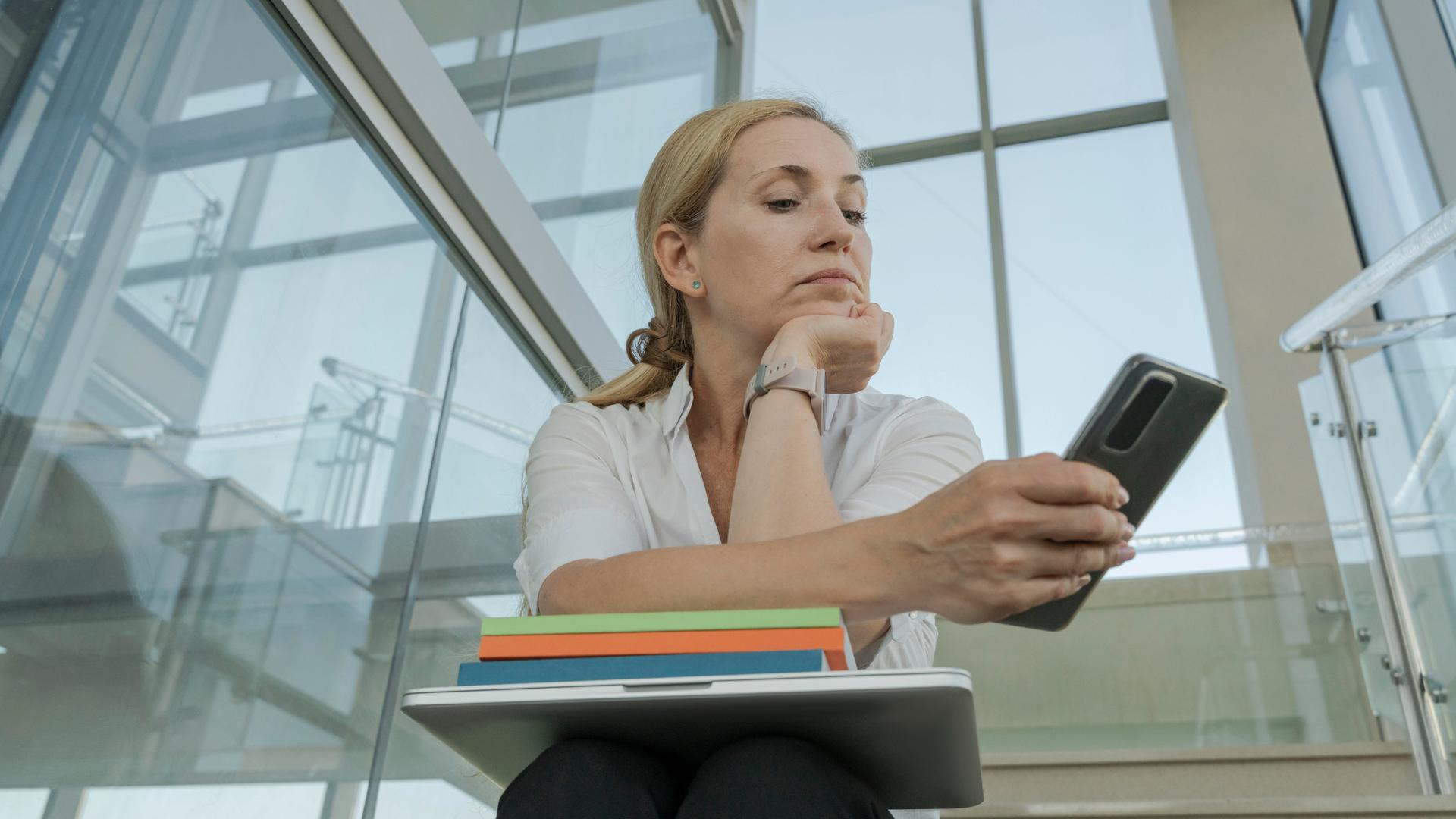 Eine Frau sitzt mit einem zugeklapptem Laptop und Büchern auf dem Schoß auf einer Treppe und schaut auf ihr Smartphone.