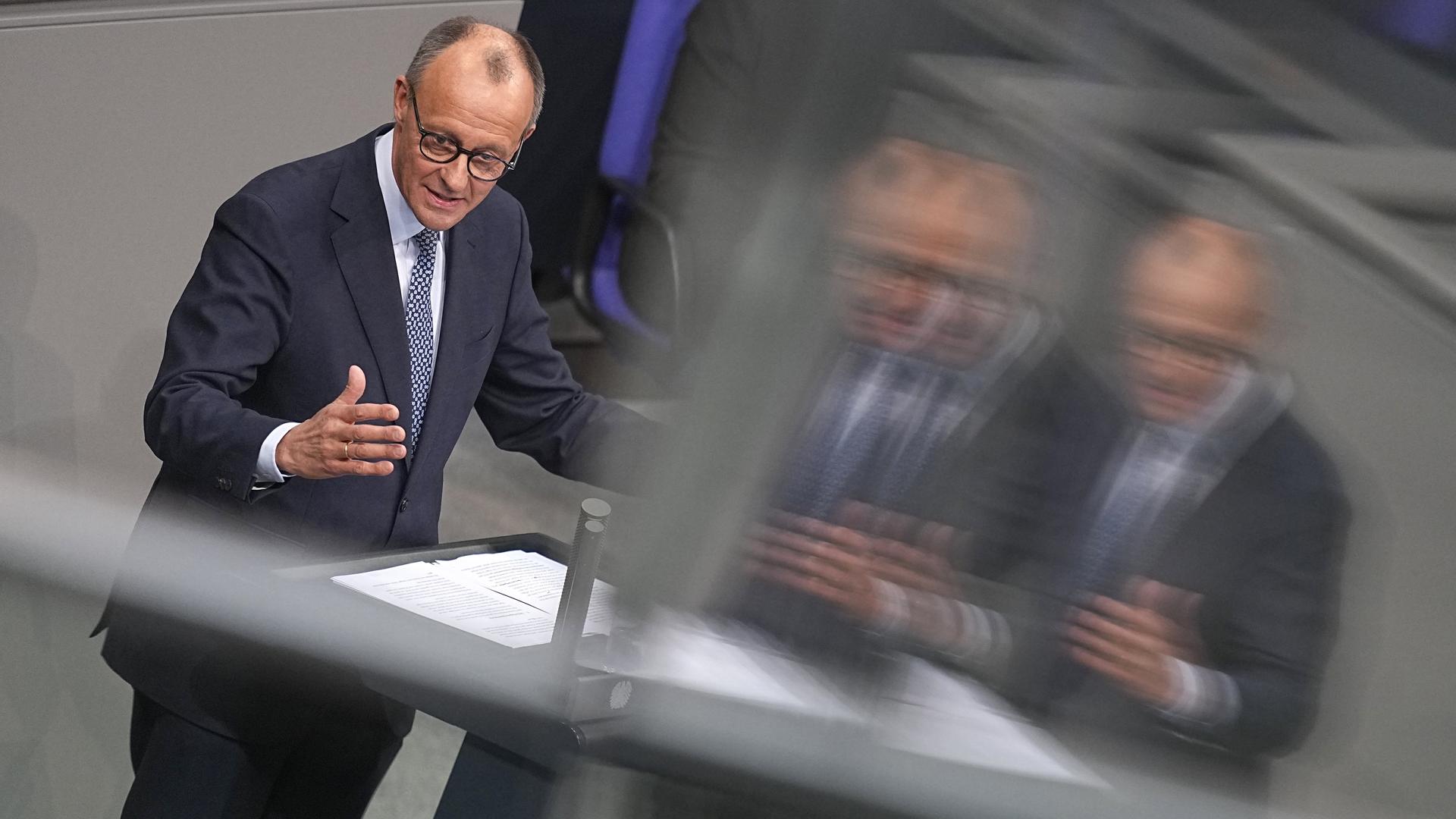 Friedrich Merz, CDU-Vorsitzender und Vorsitzender der CDU/CSU-Bundestagsfraktion, steht im Bundestag am Rednerpult. Er spiegelt sich etwas verschwommen in einer Glasscheibe.