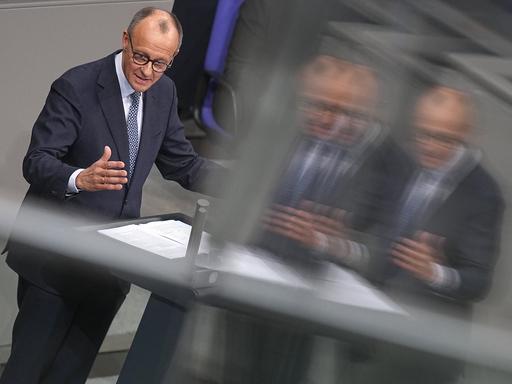 Friedrich Merz, CDU-Vorsitzender und Vorsitzender der CDU/CSU-Bundestagsfraktion, steht im Bundestag am Rednerpult. Er spiegelt sich etwas verschwommen in einer Glasscheibe.