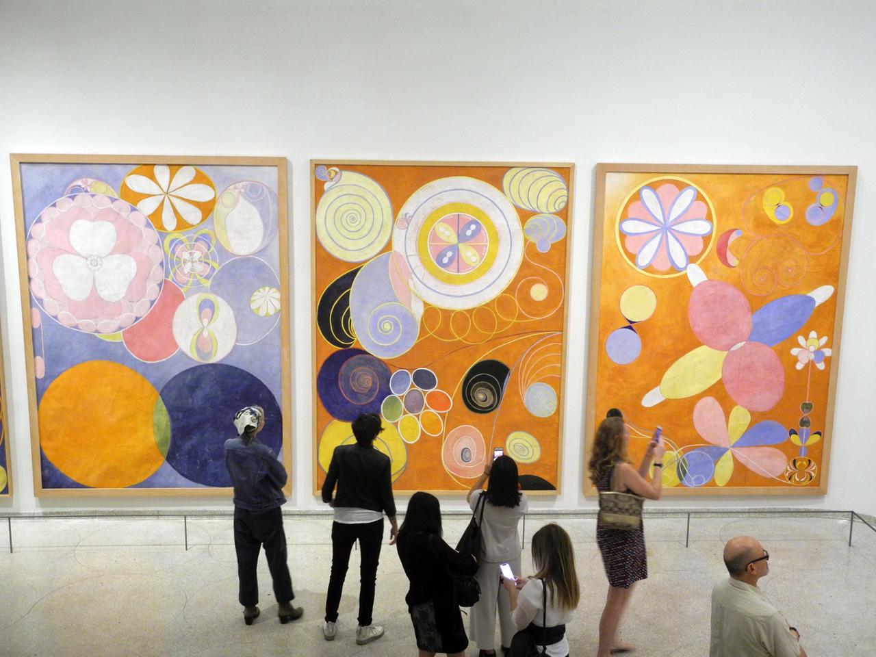In einem Museum schauen sich Besucher drei große Gemälde der schwedischen Malerin Hilma af Klint an. Es sind abstrakte Bilder.