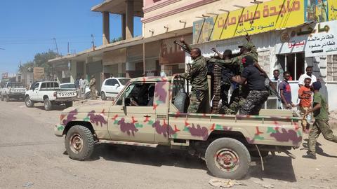 Mehrere bewaffnete Soldaten in Uniform stehen auf der Ladefläche eines Pickup. Im Hintergrund Häuser und Passanten.