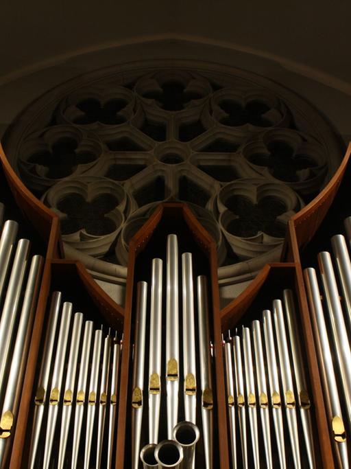 Blick auf eine große Orgel, deren Pfeifen den Blick auf ein rundes Kirchenfenster freigibt.