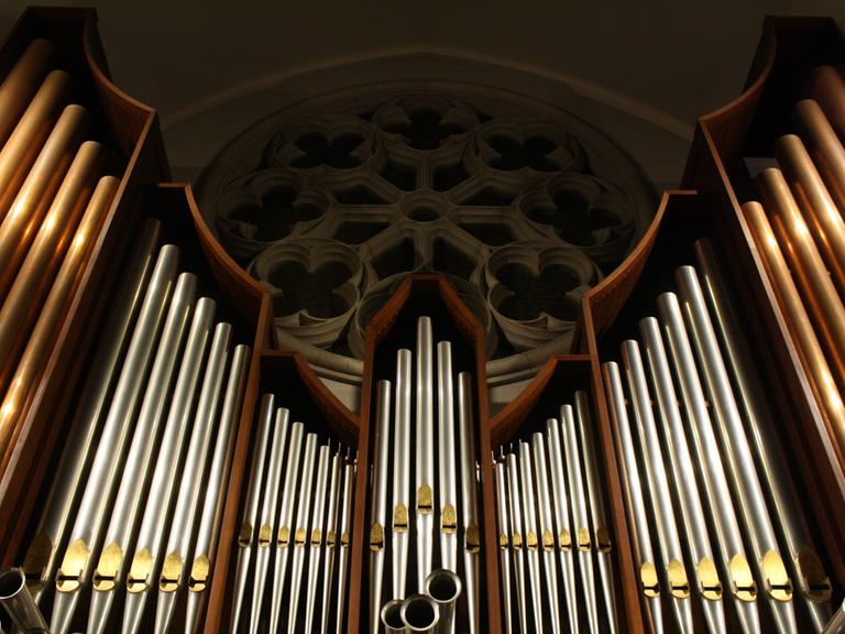 Blick auf eine große Orgel, deren Pfeifen den Blick auf ein rundes Kirchenfenster freigibt.